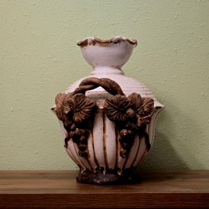 White ceramic vase with a rustic floral motif styled on a wooden shelf against a pastel wall.